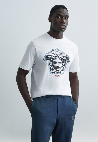Weißes Baumwoll-T-Shirt mit einem großen schwarzen und blauen Medusa-Grafik von Versace, kombiniert mit dunkelblauen Hosen mit strukturierter Oberfläche.