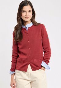 Cardigan rosso lavorato a maglia con scollatura rotonda, chiusura a bottoni e dettagli testurizzati. Camicia col celeste in vista sotto.