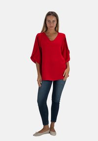 Rode V-hals blouse met brede mouwen, textuurstof en een losse pasvorm. In combinatie met donkerblauwe skinny jeans en beige ballerina's.