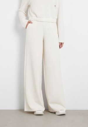 Osoba nosząca szerokie, z wysokim stanem spodnie w kolorze ecru z elastycznym pasem i bocznymi kieszeniami, w zestawie z dopasowaną bluzką z długim rękawem i białymi trampkami.