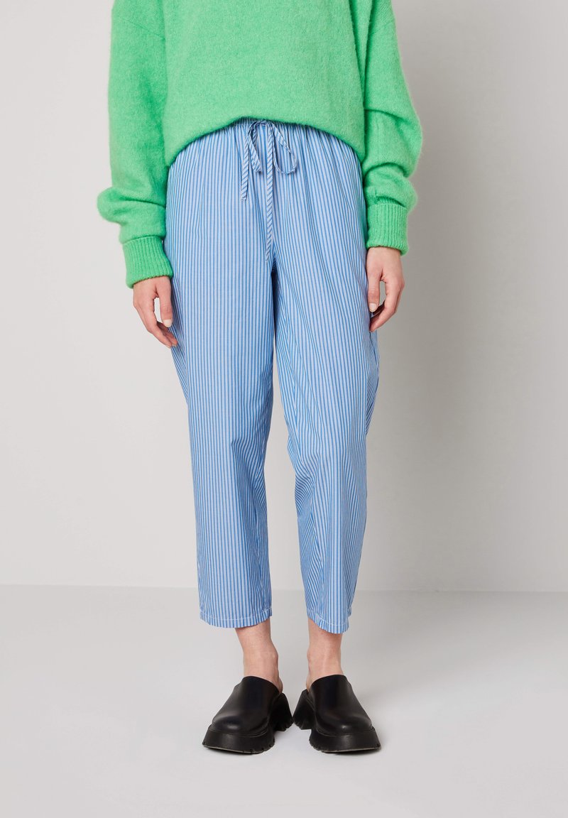 Henkilö, jolla on päällään sinivalkoiset pystysuorat raidalliset housut, joissa on kiristysnauha vyötäröllä, vihreä neulepusero ja mustat liukukengät, seisomassa valkoisella lattialla.