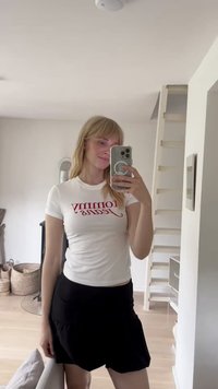 Camiseta blanca de manga corta con el logo rojo de "Tommy Jeans", combinada con una falda negra con volantes, en un espacio interior bien iluminado con suelo de madera.