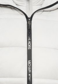Veste matelassée blanche avec ruban de fermeture éclair noir portant le texte "MICHAEL KORS", et une tirette de fermeture éclair fermée au centre avant.