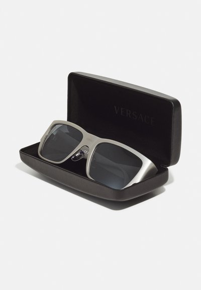 Ochelari de soare Versace într-un capac negru. Ramele sunt din metal argintiu cu lentile gri colorate și au o formă rectangulară. Detalii notabile cu logo-ul sunt prezente.
