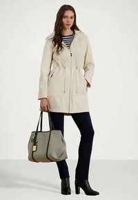 Mujer vestida con abrigo beige con capucha, pantalones azul marino, botas de tacón negras, sosteniendo un gran bolso tote gris.