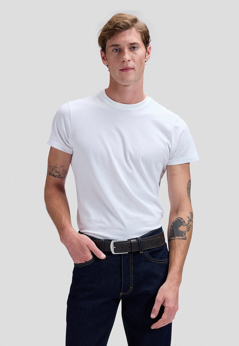 Mann mit hellbraunen Haaren, der ein schlichtes weißes T-Shirt und dunkle Jeans mit schwarzem Gürtel trägt, steht vor einem schlichten grauen Hintergrund.