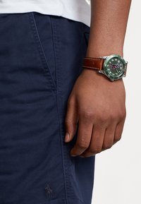 Rund klocka med grön urtavla, silverfärgad boett och brun läderrem som visas på en hand som bär marinblå shorts med en logotyp.