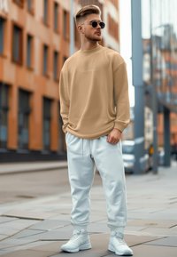 Felpa beige con logo ricamato, abbinata a pantaloni jogger bianchi. L'outfit è completato da scarpe da ginnastica bianche e occhiali da sole rotondi.