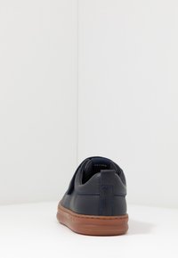 Jūras spēku zilo ādas sporta apavu ar zemu siluetu, brūnu gumijas zoli un aizmugurējo cilpu. Iezīmējas ar minimālu šuvju skaitu un gludu tekstūru.