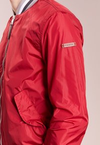 Uomo che indossa una giacca bomber rossa brillante con colletto a righe, chiusura con zip e tasca a pattina su sfondo rosa chiaro.