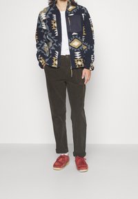 Fleecová bunda s geometrickým vícebarevným vzorem, tmavě modrým základem, předním zipem a kapsou na hrudi; doplněná tmavě hnědými kalhotami a červenými botami.