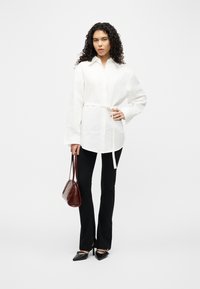 Γυναίκα με σκούρα σγουρά μαλλιά, που φοράει λευκό πουκάμισο με ζώνη, μαύρο παντελόνι, μαύρα ψηλοτάκουνα και κρατά μια σκούρα καφέ τσάντα, μπροστά σε απλό φόντο.