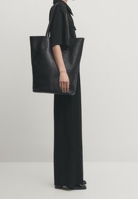 Zwarte leren tote bag met een gladde textuur, grote vorm, lange hengsels en subtiele naaddetails, vastgehouden door een persoon in zwarte kleding.