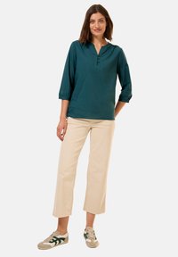 Blusa di cotone color teal con scollo a V e maniche a tre quarti, abbinata a pantaloni larghi color crema e sneakers grigie con accenti verdi.