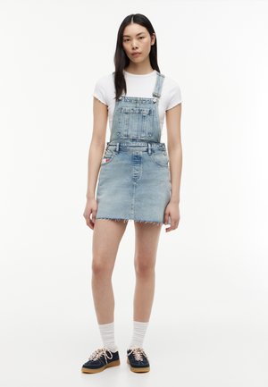 Ung kvinde med langt mørkt hår iført en hvid t-shirt, lyseblåt denim-overallsnederdel, hvide sokker og marineblå sneakers med gummisåler.
