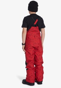 Röda snöhängselbyxor med svarta grafiska mönster, justerbara remmar och en avslappnad passform, ihop med en svart tröja och en svart mössa.