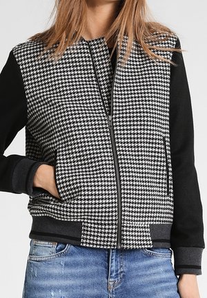 Donna che indossa una giacca con zip pied de gallo bianca e nera con maniche nere e jeans blu consumati, mano in tasca.