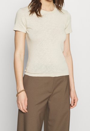 Woman wearing a fitted short-sleeve cream shirt with round neck and brown high-waist pants, accessorized with a gold chain bracelet.