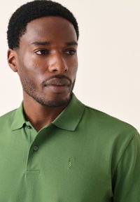 Zöld póló texturált felülettel, gombos gallérral és egy kis hímzett mintával a bal mellrészen.