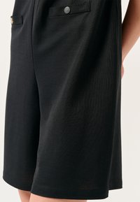 Zwarte shorts gemaakt van een gladde stof met een subtiele textuur. Heeft twee voorzakken met metalen bevestigingen en een relaxte pasvorm.