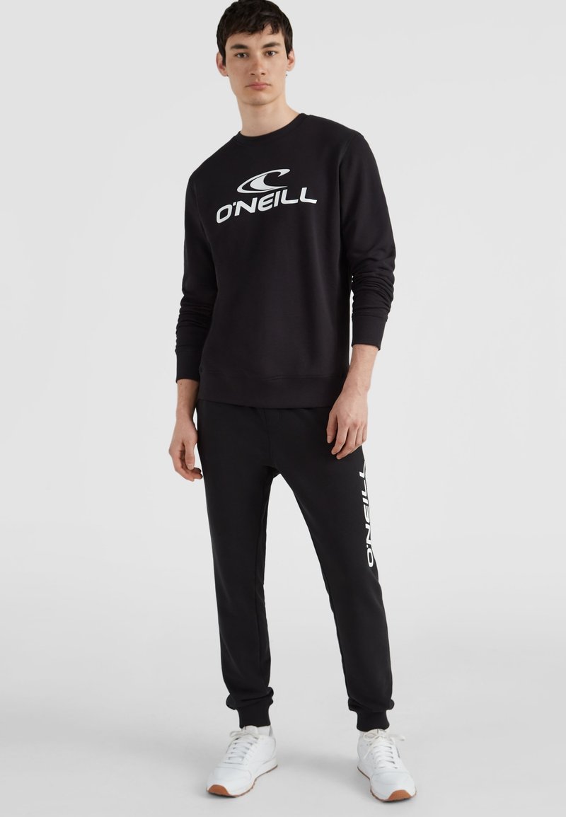 Joven de pie vistiendo una sudadera negra de O'Neill y pantalones negros a juego con zapatillas blancas, sobre un fondo claro y liso.