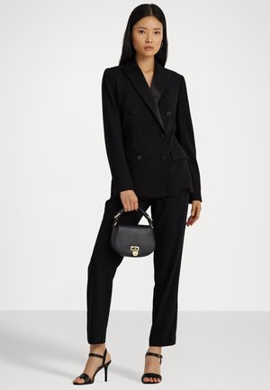 Femme portant un blazer noir croisé et un pantalon, tenant un petit sac à main noir et portant des sandales à talons noires.