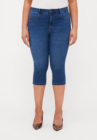 Jeans capri in denim blu scuro con vita a media altezza, tasche frontali e orlo che termina sotto il ginocchio. La modella indossa tacchi alti con stampa animale.