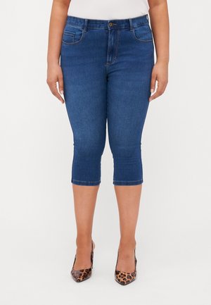 Jean capri en denim bleu foncé avec une taille mi-haute, poches avant et ourlet se terminant sous le genou. Le modèle porte des escarpins à imprimé animal.