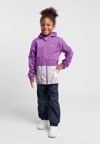 Paarse waterdichte jas met grijze onderkant, ritssluiting aan de voorkant en capuchon, gecombineerd met marineblauwe waterdichte broek en witte sneakers.