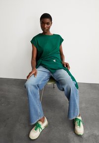 T-shirt vert à manches courtes avec un détail de nœud sur le côté, associé à un jean large bleu clair et des baskets blanches avec des accents verts.