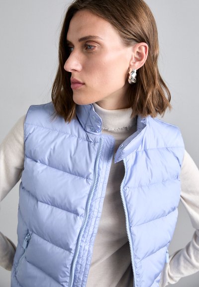 Helles blauer Puffer-Weste mit vertikaler Steppung, Druckknopf- und Reißverschlussverschluss sowie hohem Kragen. Über einem beigen Pullover getragen.