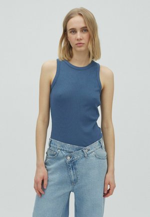 Jeune femme aux cheveux blonds mi-longs portant un haut bleu sans manches et un jean taille haute bleu clair avec une taille asymétrique.
