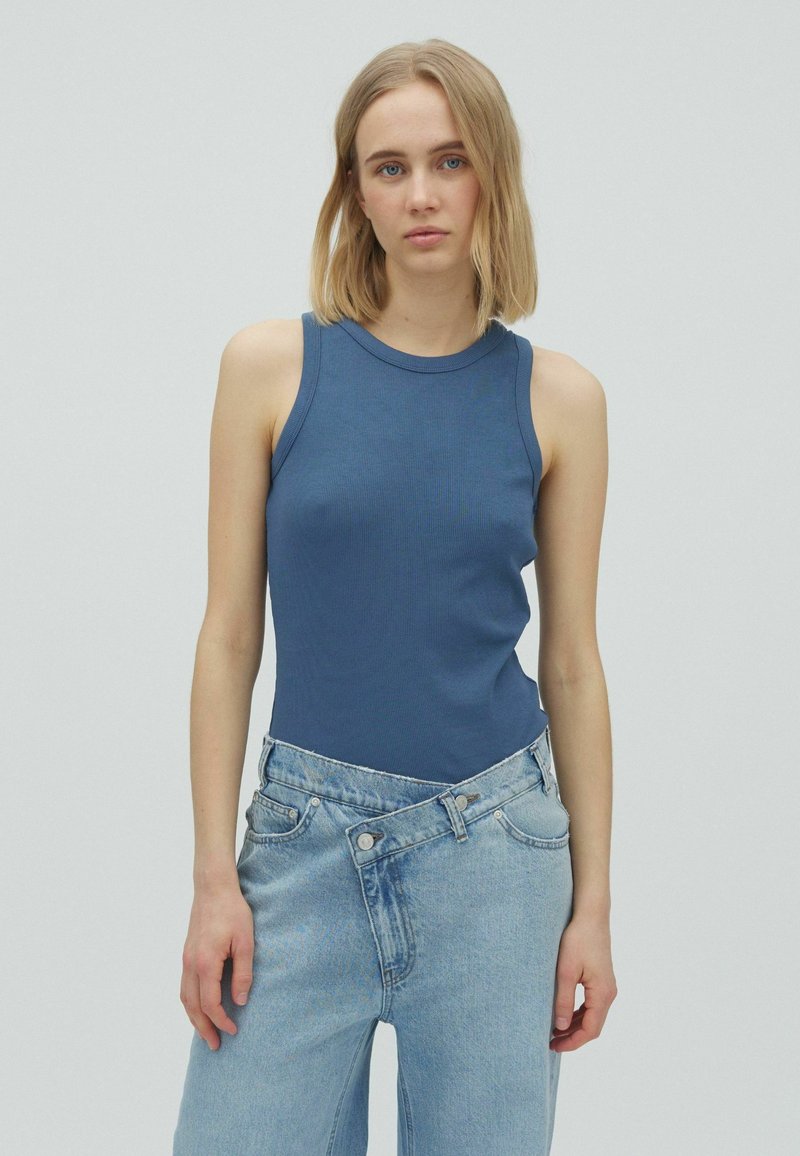 Jeune femme aux cheveux blonds mi-longs portant un haut bleu sans manches et un jean taille haute bleu clair avec une taille asymétrique.