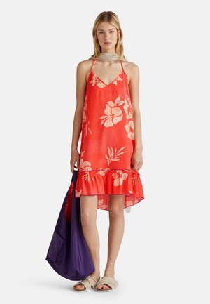 Ärmelloses rotes Kleid mit weißem Blumenmuster, Rüschensaum und V-Ausschnitt. Das Modell hält eine lila Tasche und trägt leichte Sandalen.