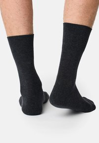 Dunkelgraue Socken werden an den Beinen getragen, sichtbar von den Knöcheln abwärts, vor einem schlichten weißen Hintergrund. Die strukturierten Bündchen sind deutlich zu sehen.