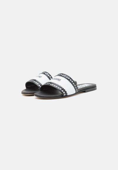 Des sandales à glisser noires et blanches avec une semelle plate, une large sangle à accents en dentelle et "Moschino" imprimé sur le dessus.