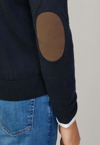 Marinblå tröja med bruna armbågsflickor, ribbade muddar och nederkant, tillverkad av mjukt material. Bärs med blå denimjeans.