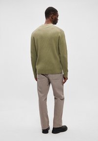 Olivovo zelený rebrovaný sveter s okrúhlym výstrihom, kombinovaný so svetlobežovými nohavicami a čiernou obuvou. Zadný pohľad, hladká textura, žiadne vzory.