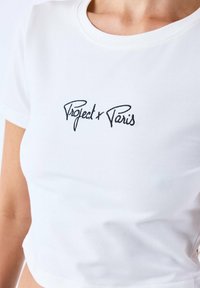 T-shirt en coton blanc avec un col rond et des manches courtes, présentant un texte noir brodé "Project x Paris" sur le devant.