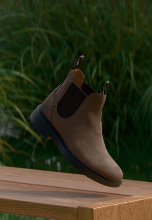 Bottine Chelsea en daim marron avec côtés élastiques noirs flottant au-dessus d'une surface en bois, feuillage vert flou en arrière-plan.