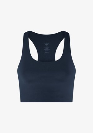 Küllasteblondide lokkis juustega naine kannab tumelilla sportrinnahoidjat, musti pükse ja valgeid tossusid, seisab ühetaustal heleda tausta ees.