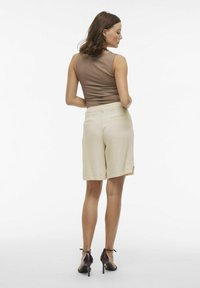 Top senza maniche color tan abbinato a pantaloni corti crema fino al ginocchio. I pantaloni corti presentano una texture liscia e tasche posteriori. La modella indossa scarpe con tacco scuro.
