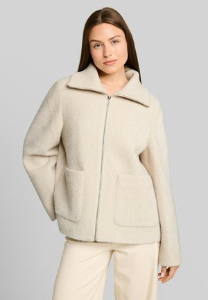 Frau mit glattem braunem Haar, die eine cremefarbene Jacke mit Reißverschluss, großem Kragen und Taschen trägt, kombiniert mit hellen Hosen, vor grauem Hintergrund.