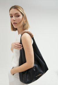 Sac en cuir noir avec une anse tressée couleur tan, affichant une forme décontractée et un accent logo doré en bas à droite.