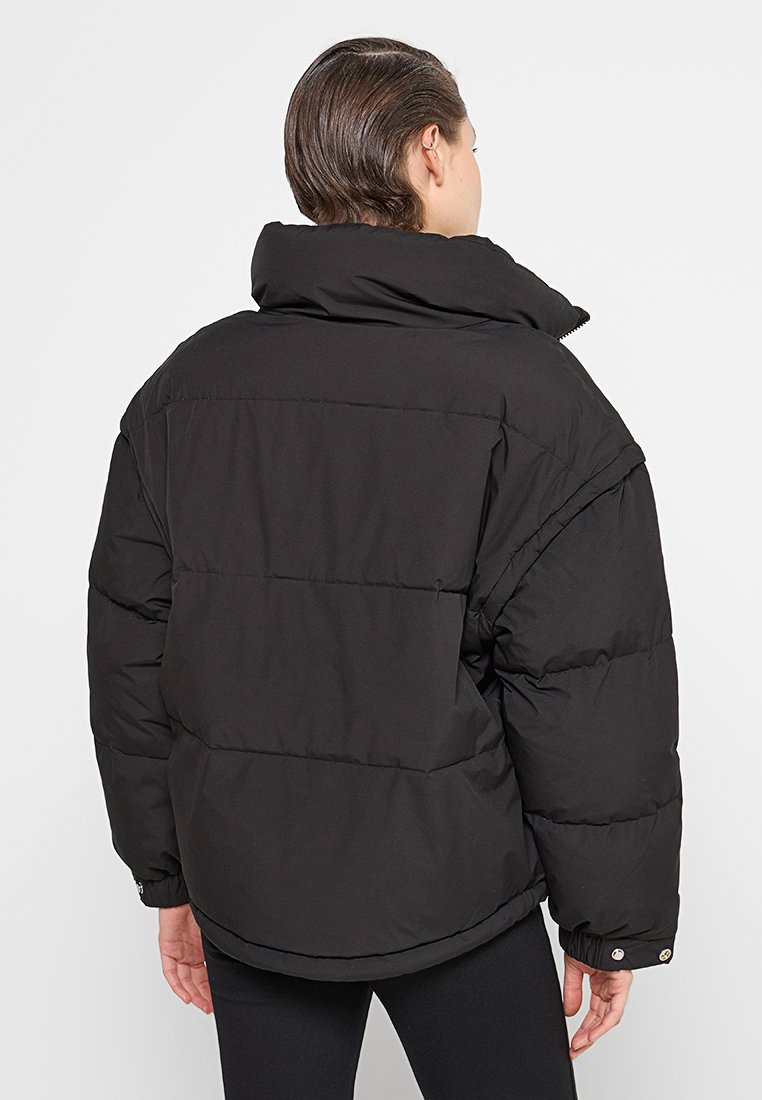 Person mit kurzem Haar, der eine schwarze Pufferjacke mit hohem Kragen trägt und mit dem Rücken zu einem schlichten Hintergrund steht.