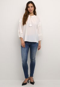Camicia bianca leggera con maniche a palloncino, scollatura con cordoncino e dettagli con nappe. Abbinata a jeans skinny blu e tacchi neri.
