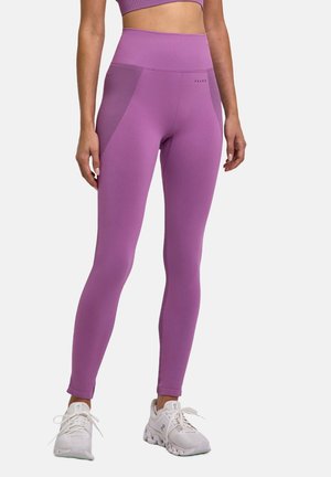 Femme portant des leggings violets taille haute et un crop top assorti, se tenant avec les mains détendues, chaussures de course blanches visibles.