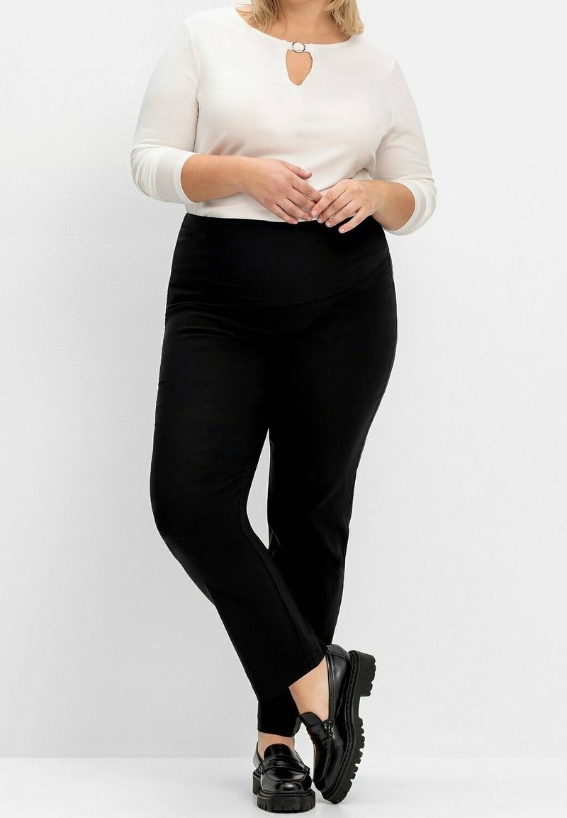 Haut blanc à manches longues avec un col à ouverture clé, associé à un pantalon noir ajusté et des chaussures noires épaisses. Textures lisses et design moderne.