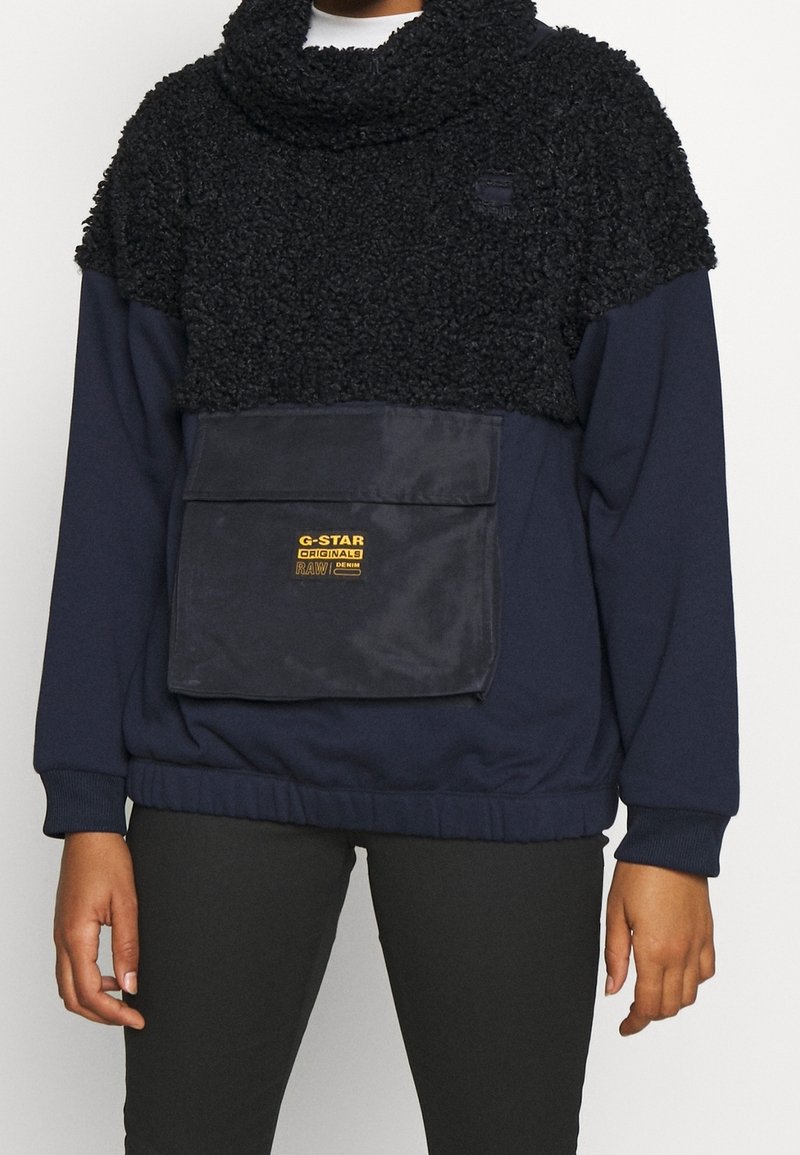 Mörkblå pullover med en svart, fluffig, texturerad övre del. Har en stor framficka med en gul logo och en hög krage.