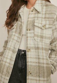 Geruite overhemdjas van zachte, gestructureerde stof. Voornamelijk crème met grijze accenten, voorzien van twee borstzakken en knoopsluitingen.